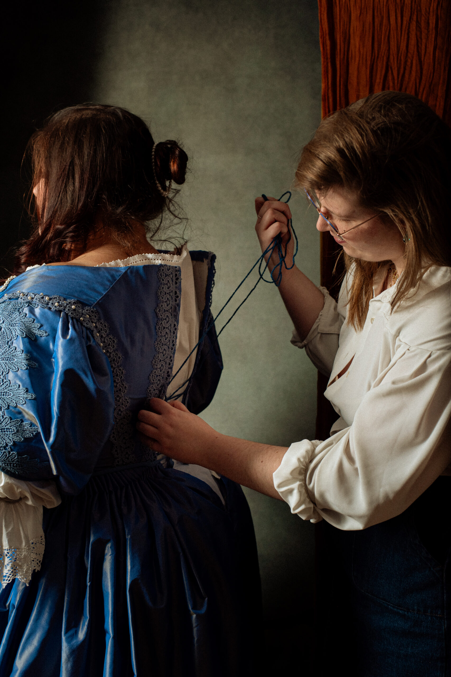 Deux femmes, dont l'une est vue de dos, s'habillent d'une robe bleue à manches longues avec de la dentelle blanche et bleue, et de volants. La seconde femme lace le dos de la robe de la première femme. La scène est éclairée par la gauche et les deux femmes sont partiellement éclairées, le côté droit de l'image étant plus sombre.